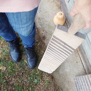 May include: A wooden washboard with a worn, rustic look. The washboard has raised ridges for scrubbing clothes. The washboard is held by a hand wearing a pink sweater and blue jeans.