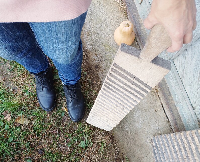 May include: A wooden washboard with a worn, rustic look. The washboard has raised ridges for scrubbing clothes. The washboard is held by a hand wearing a pink sweater and blue jeans.