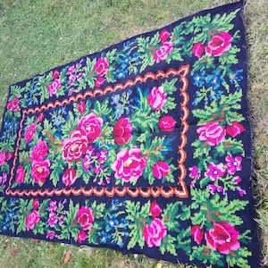 Op de afbeelding: Een zwart wollen tapijt met een bloemenpatroon in tinten roze, groen en blauw. Het tapijt heeft een rand met een gegolfde zoom.
