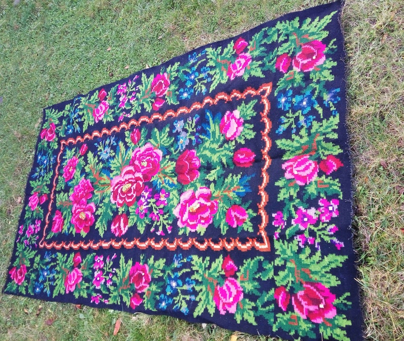 Op de afbeelding: Een zwart wollen tapijt met een bloemenpatroon in tinten roze, groen en blauw. Het tapijt heeft een rand met een gegolfde zoom.