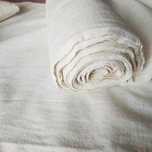 May include: A roll of white cotton fabric, unbleached and natural, is lying on a flat surface. The fabric is folded and rolled neatly, showing the texture of the material.