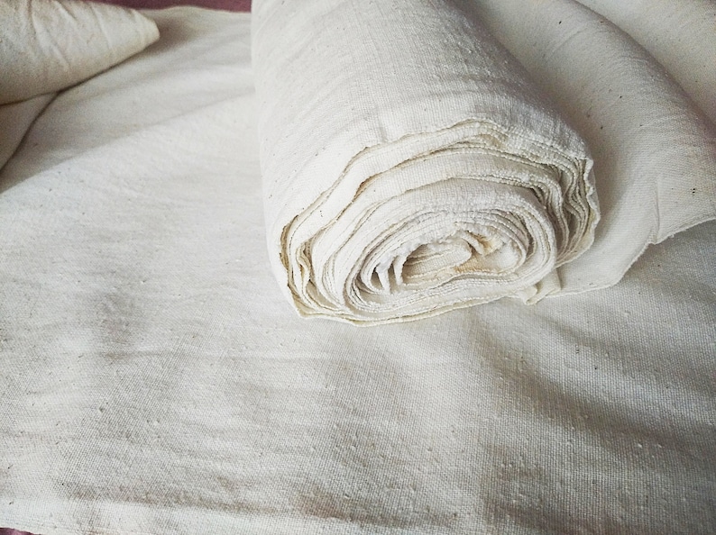May include: A roll of white cotton fabric, unbleached and natural, is lying on a flat surface. The fabric is folded and rolled neatly, showing the texture of the material.