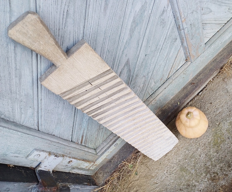 May include: A vintage wooden washboard with a light brown color and a worn surface. The washboard has a series of raised ridges for scrubbing clothes. The washboard is leaning against a weathered wooden door.