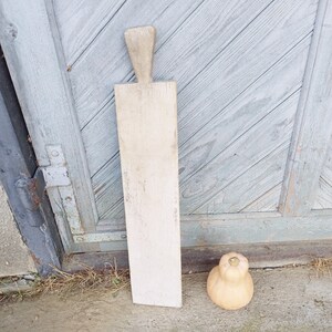 May include: A long, rectangular wooden cutting board with a handle at the top. The board is light brown and has a worn, rustic look. A small, round, light brown gourd sits on the ground in front of the board.