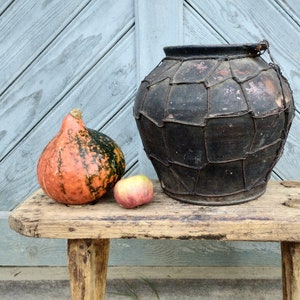 May include: A rustic wooden bench with a large, dark brown clay pot with a woven wire design, a small orange and green gourd, and a red apple.