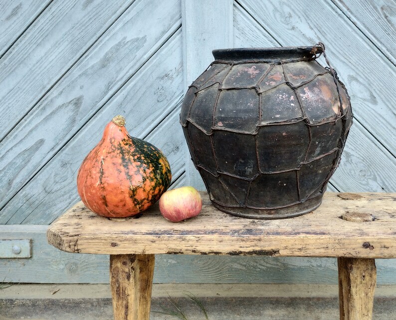 May include: A rustic wooden bench with a large, dark brown clay pot with a woven wire design, a small orange and green gourd, and a red apple.