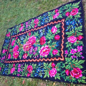 Op de afbeelding: Een zwart wollen vloerkleed met een bloemenpatroon. Het vloerkleed heeft roze, rode, groene en blauwe bloemen op een zwarte achtergrond. Het vloerkleed heeft een zwarte en bruine rand.