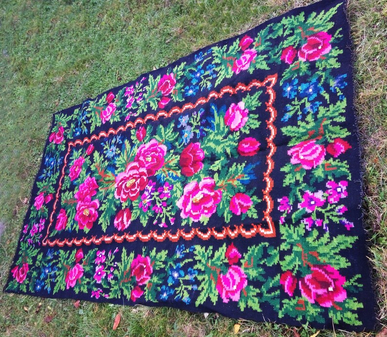 Op de afbeelding: Een zwart wollen vloerkleed met een bloemenpatroon. Het vloerkleed heeft roze, rode, groene en blauwe bloemen op een zwarte achtergrond. Het vloerkleed heeft een zwarte en bruine rand.