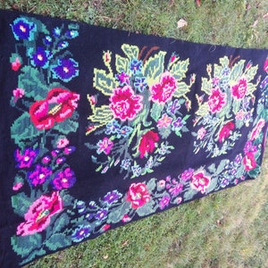 May include: A black wool rug with a floral design. The rug features pink, red, blue, and green flowers on a black background. The rug has a fringe border.