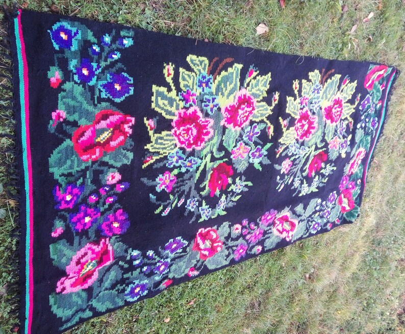 May include: A black wool rug with a floral design. The rug features pink, red, blue, and green flowers on a black background. The rug has a fringe border.