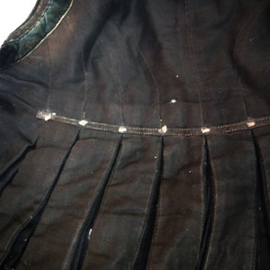 May include: Black skirt with a pleated design and a black velvet trim. The skirt has a waistband with a row of white buttons.