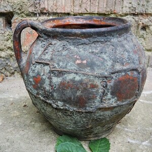 May include: A large, rustic, terracotta pot with a handle. The pot is weathered and has a dark brown and green patina. It is sitting on a concrete surface with a green leaf in the foreground.