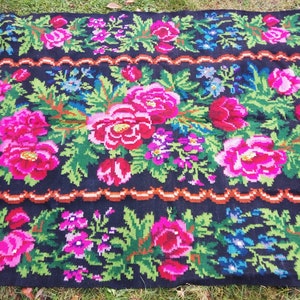 Op de afbeelding: Een zwart wollen vloerkleed met een bloemenpatroon. Het vloerkleed heeft roze, rode, groene en blauwe bloemen op een zwarte achtergrond. Het vloerkleed heeft een bruine rand.