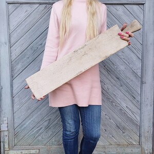 May include: A long, rectangular wooden cutting board with a light brown color and a rough, textured surface. The board is held by a person wearing a pink sweater and blue jeans.