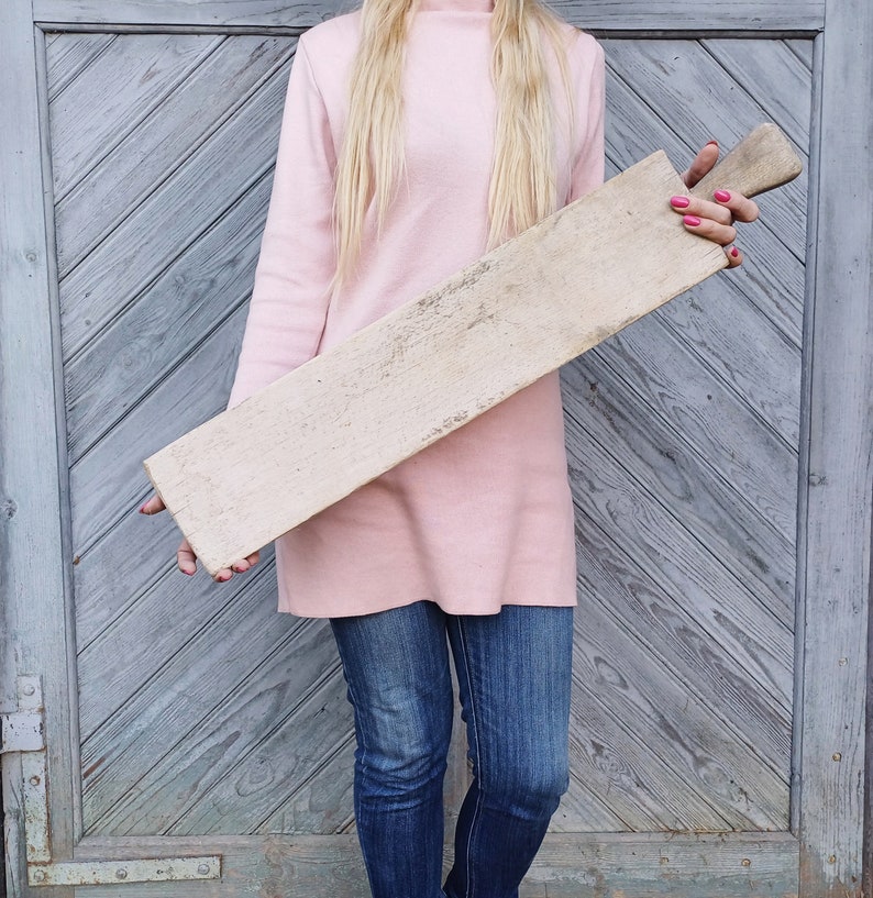 May include: A long, rectangular wooden cutting board with a light brown color and a rough, textured surface. The board is held by a person wearing a pink sweater and blue jeans.