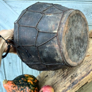 May include: A dark brown leather drum with a woven wire mesh pattern. The drum is held in a hand and has a worn, rustic appearance.