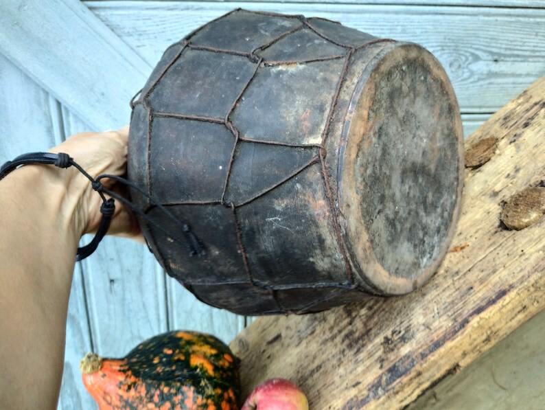 May include: A dark brown leather drum with a woven wire mesh pattern. The drum is held in a hand and has a worn, rustic appearance.