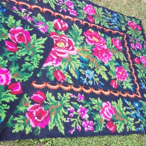 Op de afbeelding: Een zwart wollen tapijt met een bloemenpatroon met roze, rode, groene en blauwe bloemen. Het tapijt heeft een decoratieve rand.