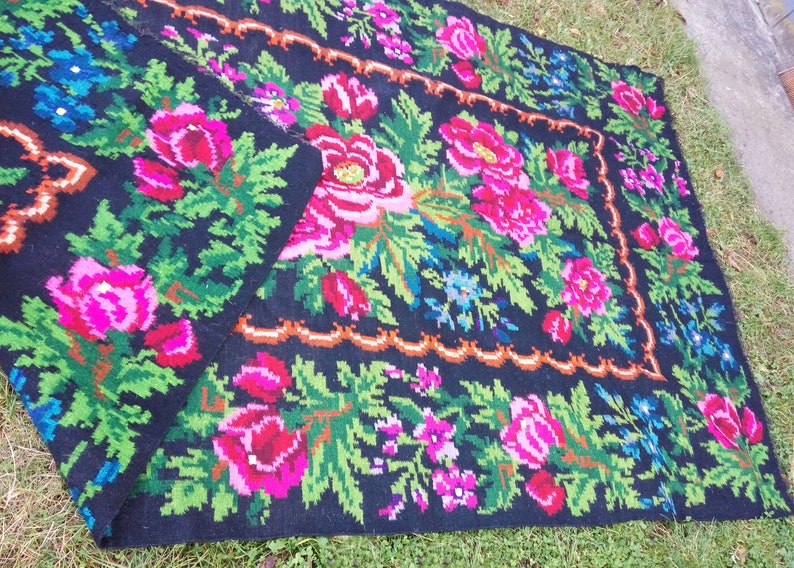 Op de afbeelding: Een zwart wollen tapijt met een bloemenpatroon met roze, rode, groene en blauwe bloemen. Het tapijt heeft een decoratieve rand.