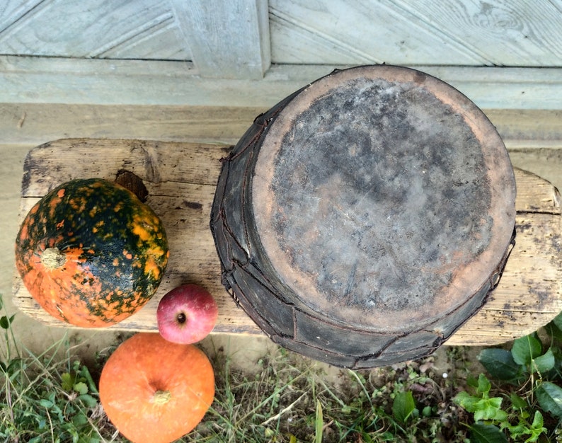 May include: A rustic, dark brown, round, woven basket with a lid sits on a wooden surface with three pumpkins. One pumpkin is orange, one is green and orange, and one is red.