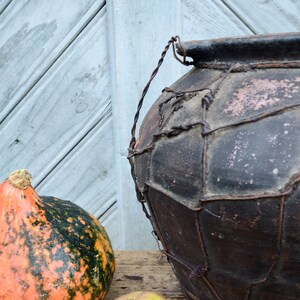 May include: A large, dark brown clay pot with a wire mesh covering. The pot has a rustic, aged appearance and is sitting on a wooden surface. A small, orange and green gourd is in the foreground.