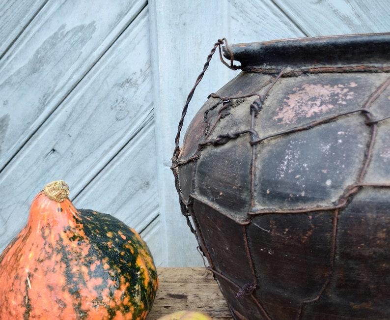May include: A large, dark brown clay pot with a wire mesh covering. The pot has a rustic, aged appearance and is sitting on a wooden surface. A small, orange and green gourd is in the foreground.