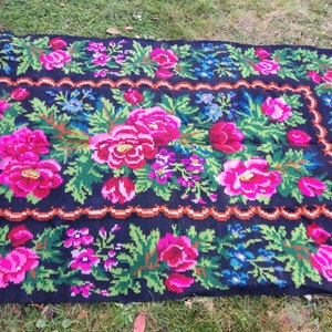 Op de afbeelding: Een traditioneel handgeweven tapijt met een bloemenpatroon. Het tapijt is zwart met roze, groene en blauwe bloemen. Het tapijt is gemaakt van wol en heeft een gefranst randje.