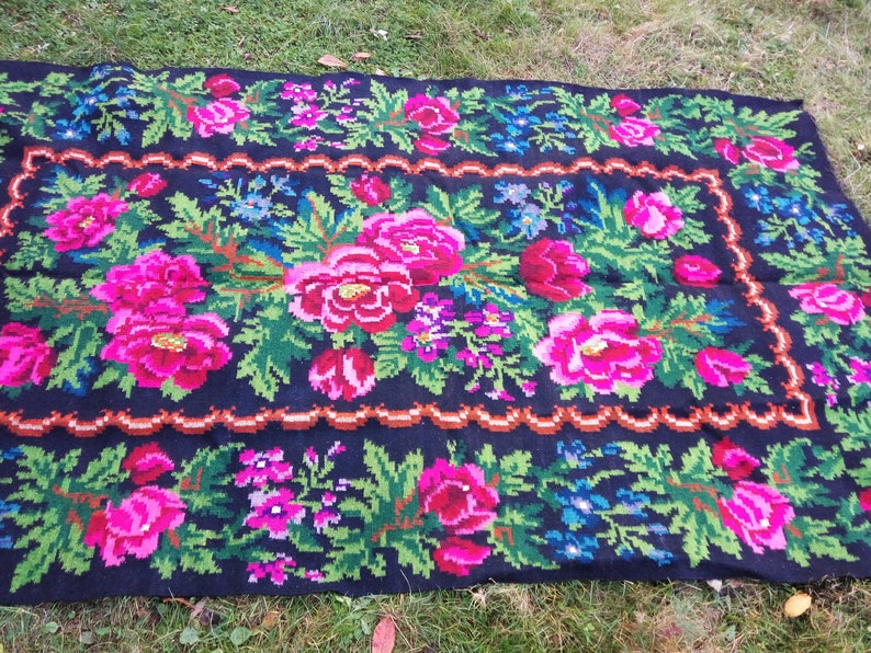 Op de afbeelding: Een traditioneel handgeweven tapijt met een bloemenpatroon. Het tapijt is zwart met roze, groene en blauwe bloemen. Het tapijt is gemaakt van wol en heeft een gefranst randje.