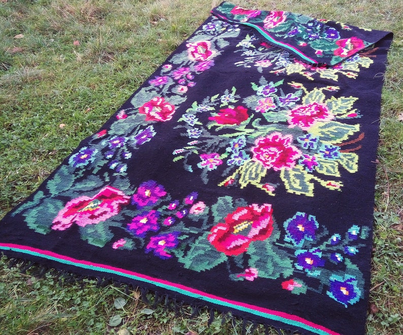 May include: A black wool rug with a floral pattern in shades of pink, red, green, blue, and purple. The rug has a fringed edge and a green and pink border.