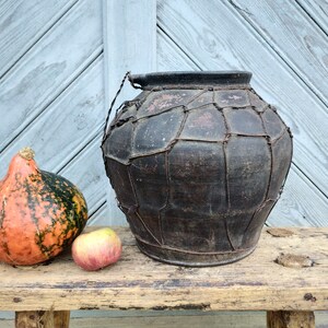May include: A large, dark brown clay pot with a woven wire exterior sits on a wooden bench. A small orange and green gourd and a red apple are in front of the pot.