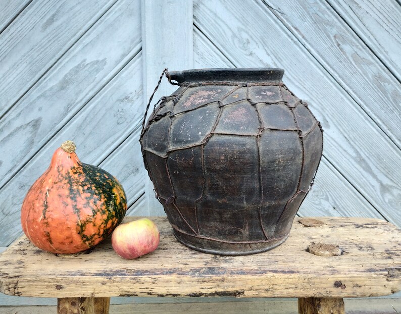 May include: A large, dark brown clay pot with a woven wire exterior sits on a wooden bench. A small orange and green gourd and a red apple are in front of the pot.