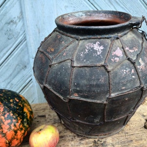 May include: A large, dark brown clay pot with a wire mesh design. The pot has a wide opening and a handle on the side. The pot is sitting on a wooden surface.