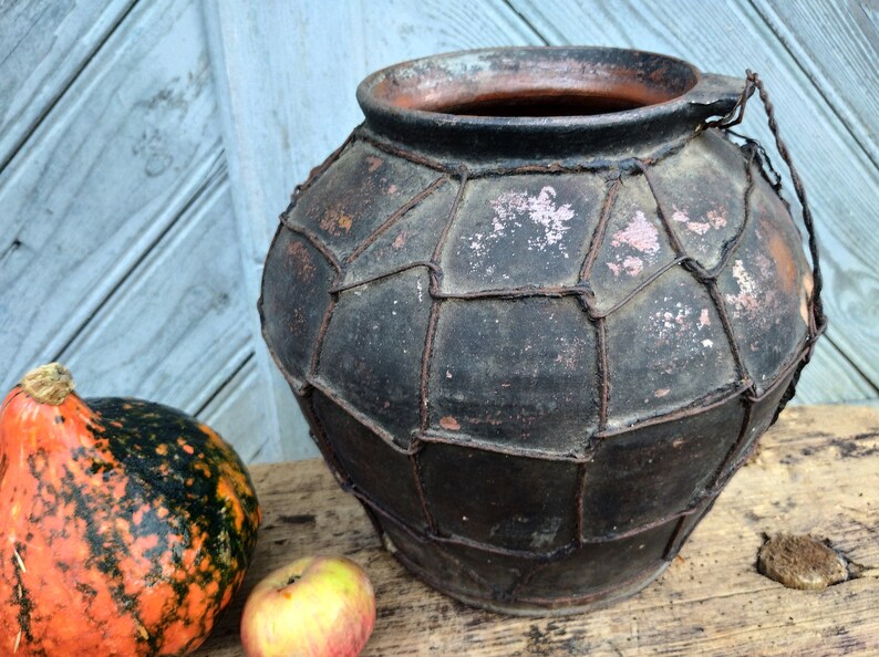 May include: A large, dark brown clay pot with a wire mesh design. The pot has a wide opening and a handle on the side. The pot is sitting on a wooden surface.