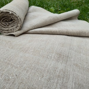 May include: A roll of natural linen fabric, partially unrolled, with a textured, neutral colour. The fabric is lying on green grass, suggesting a natural setting. The linen appears to be of a medium weight.