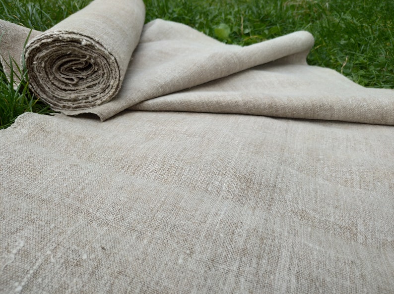 May include: A roll of natural linen fabric, partially unrolled, with a textured, neutral colour. The fabric is lying on green grass, suggesting a natural setting. The linen appears to be of a medium weight.