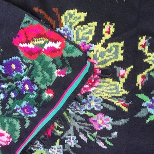 May include: A black wool rug with a colorful floral pattern. The rug features pink, red, green, blue, and yellow flowers. The rug has a fringe border.