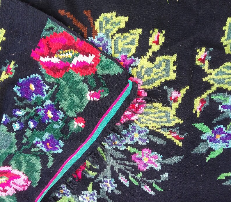 May include: A black wool rug with a colorful floral pattern. The rug features pink, red, green, blue, and yellow flowers. The rug has a fringe border.