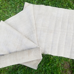 May include: A length of natural-coloured linen fabric, partly rolled and spread out on green grass. The fabric has a textured weave and a raw, unfinished edge. The neutral colour and texture suggest a rustic style.