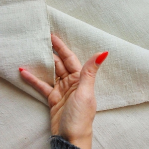 May include: A close-up of a hand holding a piece of off-white fabric. The fabric is folded in half and the hand is holding the two edges together. The fabric has a slightly textured surface.