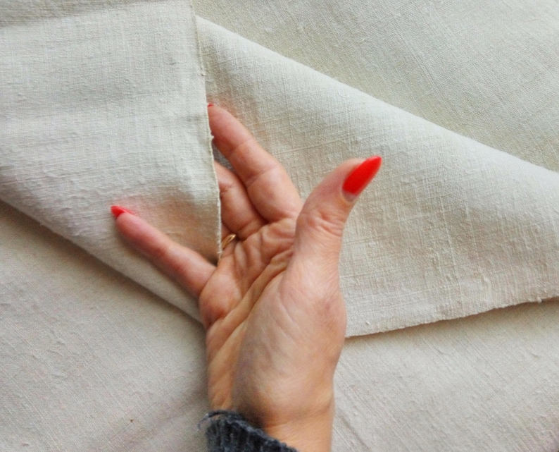 May include: A close-up of a hand holding a piece of off-white fabric. The fabric is folded in half and the hand is holding the two edges together. The fabric has a slightly textured surface.