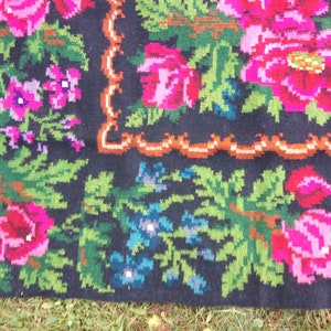 Op de afbeelding: Een close-up van een zwart wollen tapijt met een bloemenpatroon. Het tapijt heeft roze, rode, groene en blauwe bloemen op een zwarte achtergrond. Het tapijt heeft een bruine en witte rand.