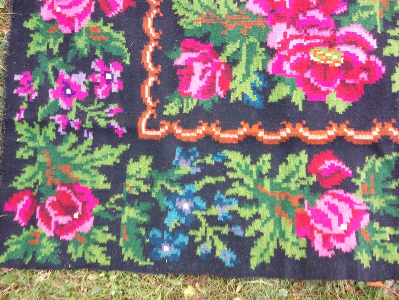 Op de afbeelding: Een close-up van een zwart wollen tapijt met een bloemenpatroon. Het tapijt heeft roze, rode, groene en blauwe bloemen op een zwarte achtergrond. Het tapijt heeft een bruine en witte rand.