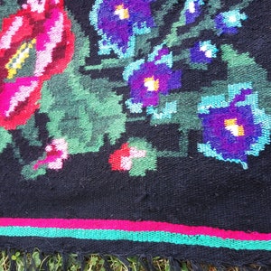 May include: A black rug with a floral pattern. The rug has red, pink, green, blue, and purple flowers. The rug has a fringe border.