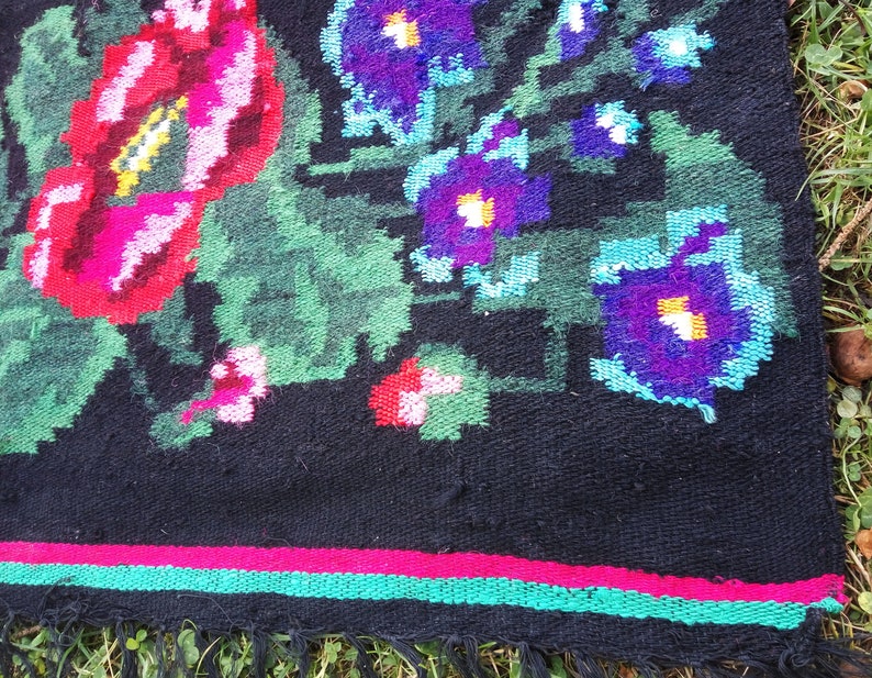 May include: A black rug with a floral pattern. The rug has red, pink, green, blue, and purple flowers. The rug has a fringe border.