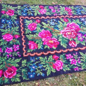 Op de afbeelding: Een zwart wollen vloerkleed met een bloemenpatroon. Het vloerkleed heeft roze, groene en blauwe bloemen op een zwarte achtergrond. Het vloerkleed heeft een rand van bruin en oranje.