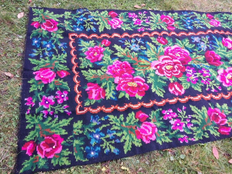 Op de afbeelding: Een zwart wollen vloerkleed met een bloemenpatroon. Het vloerkleed heeft roze, groene en blauwe bloemen op een zwarte achtergrond. Het vloerkleed heeft een rand van bruin en oranje.