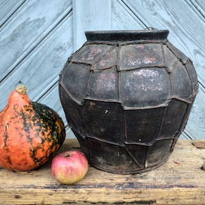 May include: A large, dark brown, ceramic pot with a woven wire design sits on a wooden surface next to a small orange and green gourd and a red apple.