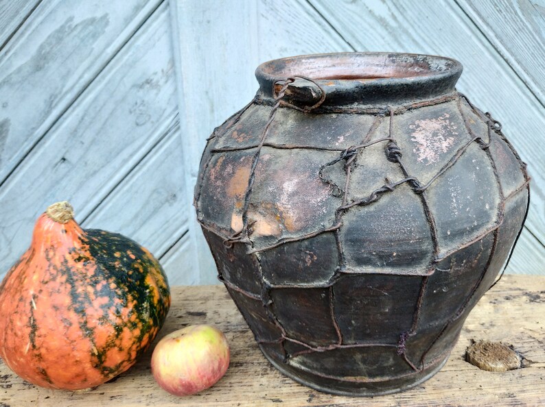 May include: A large, dark brown clay pot with a wire mesh covering. The pot has a small handle on the top. The pot is sitting on a wooden surface next to a small orange and green pumpkin and a red apple.