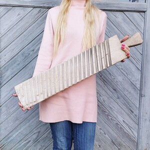 May include: A woman in a pink sweater holds a wooden washboard with a ribbed surface. The washboard is approximately 1.5 meters long.