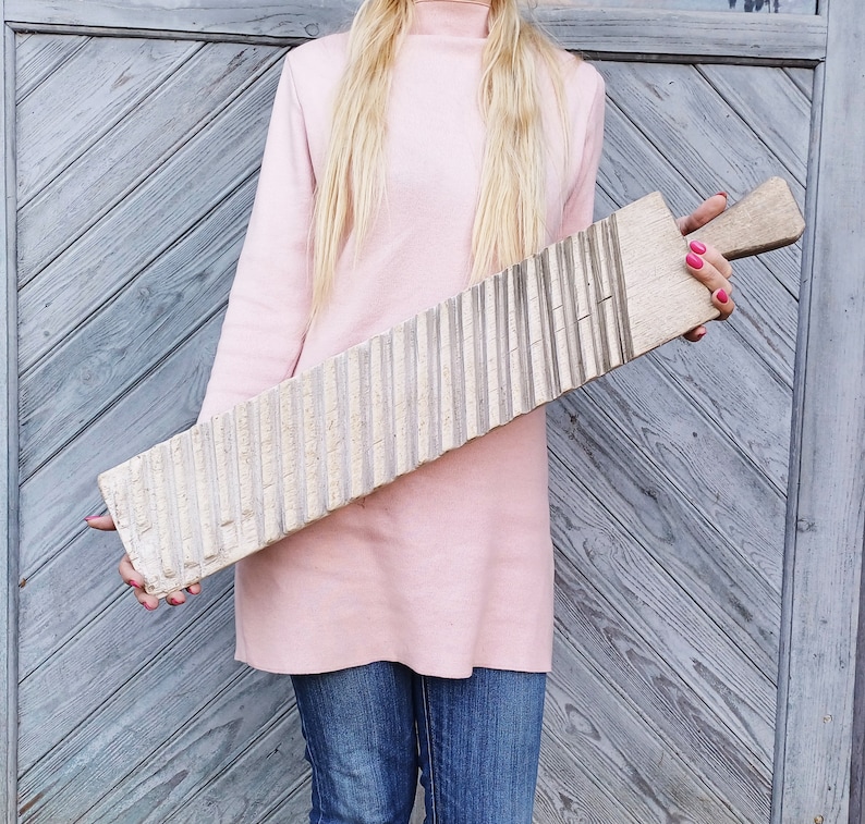 May include: A woman in a pink sweater holds a wooden washboard with a ribbed surface. The washboard is approximately 1.5 meters long.